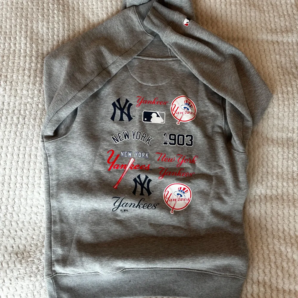 Säljer en grå hoodie från New York Yankees i storlek XL. Den har en stor logga på framsidan och flera tryck på baksidan med Yankees-tema. Perfekt för höst och vinter, och den är superbekväm med en känguruficka och justerbar huva. Nytt skick, aldrig använd. Går liten i storlek, passar mer som M/L.. Hoodies.