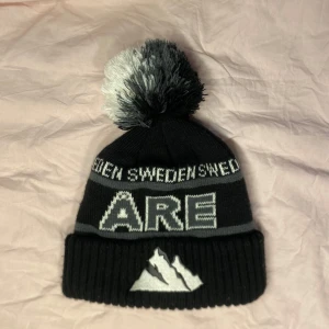 Svart mössa med tofs och text - Säljer en svart mössa med en stor, fluffig tofs i svart och grått. Mössan har texten 'ARE' och 'SWEDEN' i vitt och grått, samt ett  bergsmotiv framtill. Perfekt för kalla dagar i skidbacken eller bara för att hålla sig varm och stilren i vinter.💕
