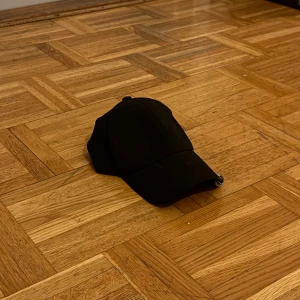 Svart keps med ringdetaljer - Säljer en stilren svart keps i mycket bra skick. Den har coola ringdetaljer på skärmen som ger en unik touch. Perfekt för både vardag och fest. Passar de flesta huvudstorlekar genom justerbar rem. Endast: 39