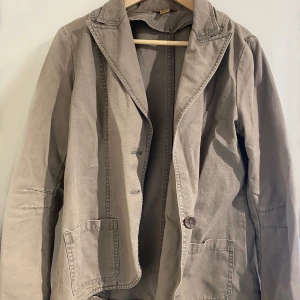 Beige kavaj - Säljer en snygg lite grön färgad  kavaj i bra skick. Den har en klassisk design med knappar framtill och två fickor. Perfekt för höst och vår, och funkar både till vardags och fest. Passar bra till jeans eller chinos. Står att den är 44 i storlek men mer som en 36. Den är felfri bara skrynklig 