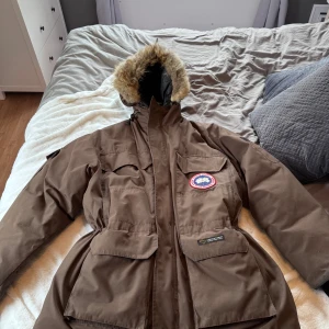 Brun parkajacka från Canada Goose - Säljer en brun parkajacka från Canada Goose i storlek L. Jackan har en mysig pälsfodrad huva och flera praktiska fickor. Perfekt för kalla vinterdagar! Den har varit med ett tag så det finns en skada på ena mudden och en skada på pälsen. 