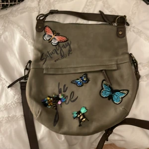 Grön axelväska med fjärilar och ros - Säljer en superfin grön axelväska med broderade fjärilar och en stor röd ros. Väskan har en cool text 'Saturday mood' och detaljer som paljetter och dragkedja. Perfekt för att ge din outfit en unik touch! 🦋🌹