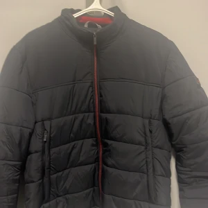 Svart pufferjacka från Superdry - Säljer en svart pufferjacka från Superdry i mycket bra skick. Jackan har en snygg röd dragkedja och praktiska fickor med dragkedjor. Perfekt för kyliga höst- och vinterdagar. Den är långärmad och har en modern stil. Passar både till vardags och lite finare tillfällen.