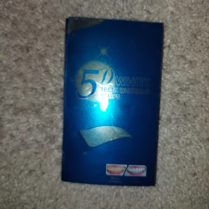 Ett paket tandblekning stripes - 5D White Teeth Whitening Strips i en blå förpackning. Innehåller flera remsor för tandblekning. Instruktioner på baksidan för enkel användning. Perfekt för att få ett vitare leende hemma.