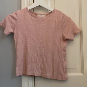 Rosa ribbad topp från H&M - Söt rosa ribbad topp från H&M i storlek 134/140. Den har korta ärmar och ett diskret mönster med små hål. Perfekt för vår och sommar! Toppen är i mycket bra skick och passar både till vardags och lite finare tillfällen.