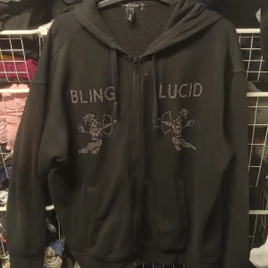 Svart hoodie från Bling Lucid - Säljer en svart hoodie från Bling Lucid i storlek L. Den har en dragkedja framtill och är dekorerad med glittriga stenar som formar texten 'BLING LUCID' och två änglar. Perfekt för en cool och avslappnad stil. Passar bra till höst och vår!