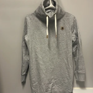 Grå låg hoodie från edc - Säljer en supermysig grå låg hoodie från edc. Den har en hög krage med snörning och ett snyggt märke på bröstet. Perfekt för kyliga dagar med sina långa ärmar och ribbade muddar. Passar både till vardags och chillkvällar. 🩶