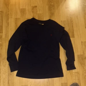 Mörkblå långärmad tröja från Polo Ralph Lauren - Säljer en snygg mörkblå långärmad tröja från Polo Ralph Lauren. Den har en klassisk design med det ikoniska röda logotypbroderiet på bröstet. Perfekt för både vardag och lite finare tillfällen. Tröjan är i mycket bra skick och gjord av mjukt bomullsmaterial. Använt max två gånger 