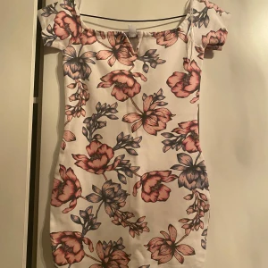Blommig klänning med korta ärmar - Säljer en superfin blommig klänning i vitt med rosa och blå blommor. Den har en söt v-ringning och korta ärmar, perfekt för sommarens fester. Klänningen är i mycket bra skick och har en tight passform som framhäver figuren. Perfekt för både vardag och fest!