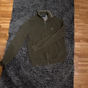 Olivgrön kofta från Gant - Säljer en snygg olivgrön kofta från Gant i mycket bra skick. Den har en dragkedja framtill och är perfekt för höst och vår. Koftan är långärmad och gjord i ett mjukt material som känns skönt mot huden. Perfekt för både vardag och lite finare tillfällen!