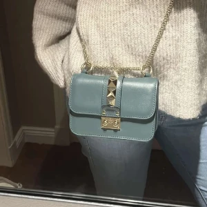 Super fin Valentino väska  - Snygg blå axelväska i skinn med guldkedja och coola nitar på locket. Perfekt för en utekväll eller när du vill lägga till lite edge till din outfit. Väskan har en praktisk storlek och en säker låsning framtill.