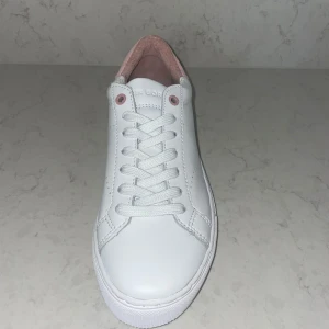 Vita sneakers med rosa detaljer - Säljer ett par snygga vita sneakers med rosa detaljer på hälen. Skorna har en klassisk snörning och är helt nya aldrig använda. Perfekta för vår och sommar, och passar till nästan alla outfits. De är bekväma och stilrena, ett måste i garderoben!