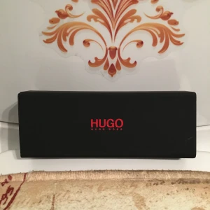 Svart glasögon från Hugo Boss - Säljer en elegant svart parfymförpackning från Hugo Boss med röd logotyp. Perfekt för den stilmedvetna som vill ha en lyxig doftupplevelse. Förpackningen är rektangulär och stilren, vilket gör den till en snygg detalj i badrummet eller på sminkbordet.