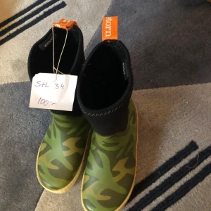 Gröna gummistövlar från Tretorn - Säljer ett par coola gröna gummistövlar från Tretorn i storlek 34. De har ett snyggt camouflage-mönster och är perfekta för regniga dagar. Stövlarna har en praktisk dragflik i orange för enkel påtagning. Perfekta för höst och vår!