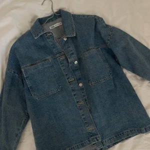 Jeansjacka från Zara - Säljer en snygg jeansjacka från Zara i storlek M. Jackan har en klassisk blå denimfärg och är utrustad med knappar framtill. Perfekt för vår och höst, med en normal passform och långa ärmar. Den är i mycket bra skick och redo för nya äventyr! Aldrig använd!!