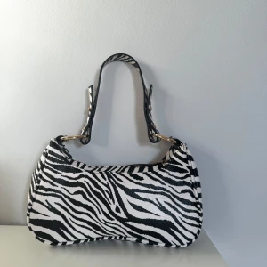 Zebramönstrad axelväska - Snygg axelväska med zebramönster i svart och vitt. Väskan har en stilren design med guldfärgade detaljer och en bekväm axelrem. Perfekt för att ge din outfit en trendig touch!