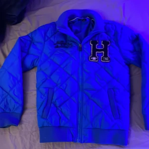 Blå quiltad jacka med bokstavsdetalj - Säljer en snygg blå quiltad jacka med dragkedja och en stor bokstav 'H' på bröstet. Jackan har långa ärmar och ribbade muddar vid ärmslut och nederkant. Perfekt för höst och vår!