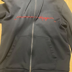Svart hoodie från Hugo Boss - Säljer en stilren svart hoodie från Hugo Boss med röd logga på bröstet. Den har en dragkedja framtill. Perfekt för både vardag och chill. Passar bra till jeans eller joggers. Hör av dig om du är intresserad! 🖤