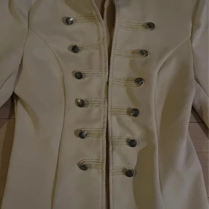 Beige kavaj från Chloé - Säljer en elegant beige kavaj från Chloé i mycket bra skick. Den har en figursydd passform med dekorativa knappar framtill. Perfekt för höst och vår, och funkar både till vardags och fest. Kavajen har långa ärmar och en stilren design. Passa på att köpa denna stiliga kavaj!