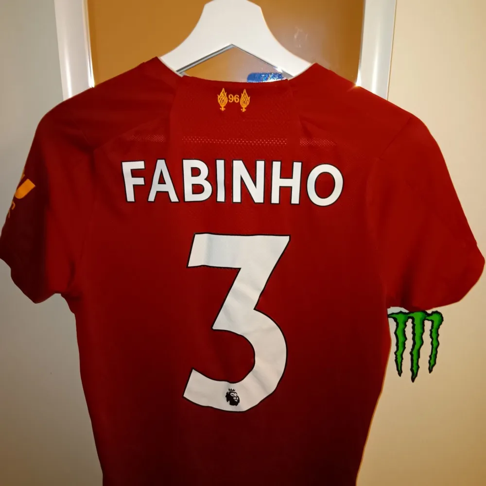 Säljer en röd Liverpool FC fotbollströja i mycket bra skick. Den är kortärmad med vita detaljer och har 'Fabinho' och nummer 3 på ryggen. Tröjan har klubbens emblem och sponsorloggor på framsidan. Perfekt för alla LFC-fans! Storlek 146. T-shirts.