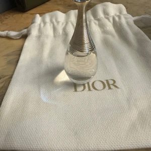 J'adore Eau de Parfum från Dior - Säljer en elegant och lyxig J'adore Eau de Parfum från Dior. Flaskan är liten och stilren med en genomskinlig design och en vacker, smal hals i silver. Perfekt för att ha med sig i väskan för en touch av glamour när som helst. Kommer med en vit påse med Dior-loggan.