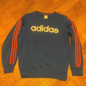 Blå tröja från Adidas - Säljer en snygg blå tröja från Adidas med de klassiska tre ränderna i rött längs ärmarna. Tröjan har en rund halsringning och är perfekt för en sportig look. Den är i bra skick och passar perfekt för höst och vår.