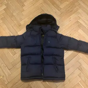 Säljer en snygg mörkblå dunjacka från Polo Ralph Lauren. Jackan har en pufferstil med långa ärmar och en praktisk huva. Perfekt för kalla vinterdagar. Den är i mycket bra skick och har en dragkedja framtill. Passar perfekt för både vardag och utomhusaktiviteter. Vi kan diskutera priset!