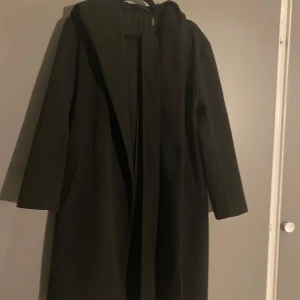 Svart kappa med huva - Säljer en stilren svart kappa med huva. Den har långa ärmar och ett knytbälte i midjan för en snygg passform. Perfekt för höst och vinter, håller dig varm och trendig. Passar både till vardags och festliga tillfällen.