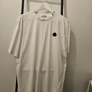 Vit t-shirt från Moncler - Säljer en stilren vit t-shirt från Moncler i bomull. Den har en liten svart logga på bröstet och Moncler-text på ryggen. Perfekt för en casual look eller att klä upp med en snygg jacka. Passar till alla säsonger och är ett måste i garderoben!