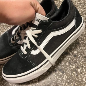 Svarta Vans sneakers - Säljer ett par klassiska svarta Vans sneakers i bra skick. De har den ikoniska vita sidostripen och vita snören . Perfekta för vardagsbruk och ger en skön streetstyle-look. Sulan är vit med det röda 'Off the Wall'-märket på hälen.