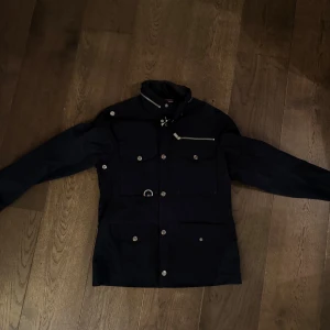 Svart jacka från Snoot - Snygg svart jacka från Snoot i storlek M. Den har en stilren design med knappar och en praktisk bröstficka. Perfekt för höst och vår, med långärmade ärmar och en klassisk krage. Passar både till vardags och lite finare tillfällen. Nyskick ca 5500
