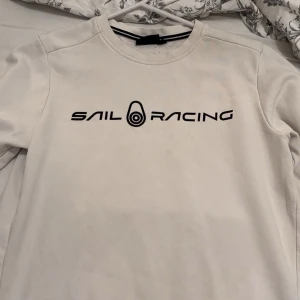 Vit tröja från Sail Racing - Säljer en stilren vit tröja från Sail Racing i mycket bra skick. Den har en enkel design med svart text på bröstet. Perfekt för en avslappnad stil eller som ett lager under jackan. Passar både till vardags och lite sportigare tillfällen. Passar XS/S