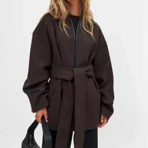 Brun kappa med bälte - Säljer en stilren brun kappa med bälte i midjan. Den har en modern, loose passform och långa ärmar. Perfekt för höst och vår, och funkar både till vardags och fest. Dragkedja framtill för en enkel look.