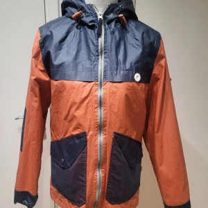 Orange och marinblå vindjacka från Bellfield - Säljer en snygg orange och marinblå vindjacka från Bellfield. Jackan har en dragkedja framtill och praktiska fickor. Perfekt för höst och vår med sin lätta och skyddande design. Den har en justerbar huva och är i mycket bra skick.
