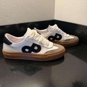 Vita sneakers med svart detalj - Säljer ett par snygga vita sneakers med svart detalj på sidan och beige mocka vid tån. De har en brun sula och vita snören. Perfekta för en casual look och passar till det mesta i garderoben.