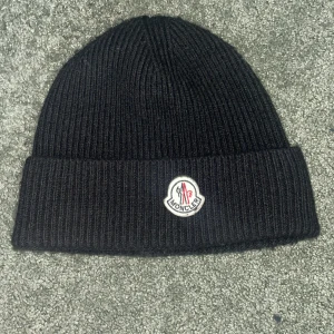 Svart ribbad mössa från Moncler.äkta moncler  - Säljer en snygg svart ribbad mössa från Moncler. Den är tillverkad i Italien av 70% ull och 30% kaninhår, vilket gör den supermjuk och varm. Perfekt för kalla vinterdagar! Mössan har Monclers ikoniska logga framtill. Den är äkta och kostar 2800kr ny pris 