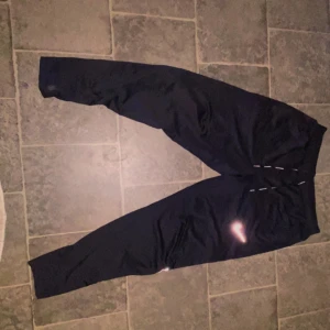 Svarta träningsbyxor från Nike - Säljer ett par svarta Nike Dri-FIT träningsbyxor i storlek M. Perfekta för löpning eller gymmet med bekväm passform och snörning i midjan. Byxorna har en liten Nike-logga och texten 'Nike Running' längs benet. De är i mycket bra skick och redo för nya äventyr! Helt ny och äkta