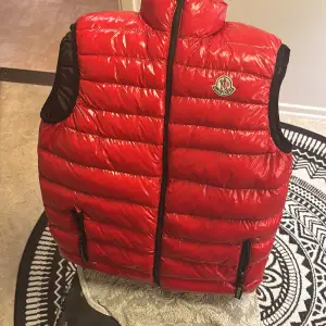 Säljer en snygg röd dunväst från Moncler i storlek S. Västen har en glansig finish med dragkedja och Moncler-logga på bröstet. Perfekt för kyliga dagar och ger en stilren look. Inuti finns en rolig serietidningsdetalj. Passar både höst och vinter.