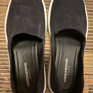 Vagabond skor - Säljer ett par snygga svarta slip-on sneakers från Vagabond. De är i bra skick och har en stilren design med vit sula. Perfekta för både vardag och lite mer avslappnade tillfällen. Skorna är bekväma och lätta att ta på och av. 