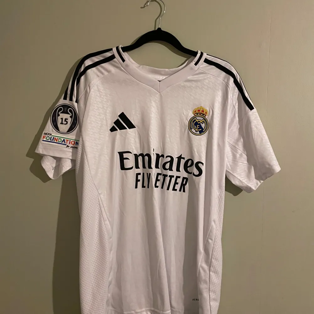 Säljer en Real Madrid fotbollströja med Vini Jr. och nummer 7 på ryggen. Tröjan är kortärmad och har svarta detaljer samt ett UEFA-märke på ärmen. Den är i slim fit och tillverkad av Adidas. Perfekt för alla fotbollsfans!. T-shirts.