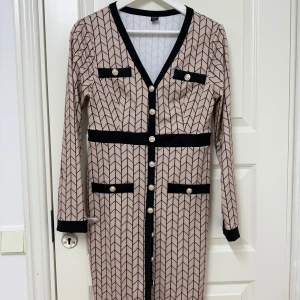 Beige och svart mönstrad klänning från Shein - Snygg beige klänning från Shein med svart mönster och detaljer. Den har en v-ringad design med knappar framtill och långa ärmar. Perfekt för höst och vår med sitt stilrena utseende. Klänningen har också dekorativa fickor och ett bälte i midjan för en elegant look. Helt ny, oanvänd och redo för en ny användare!