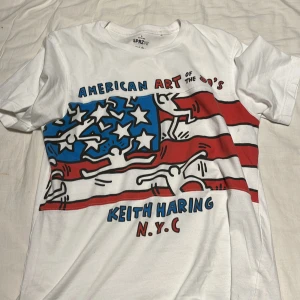 Vit uniqlo  t-shirt med Keith Haring-design - Säljer en vit t-shirt från Uniqlo med en cool Keith Haring-design. Den har ett färgglatt tryck med amerikanska flaggan och texten 'American Art of the 80's'. Perfekt för dig som gillar konst och streetwear!