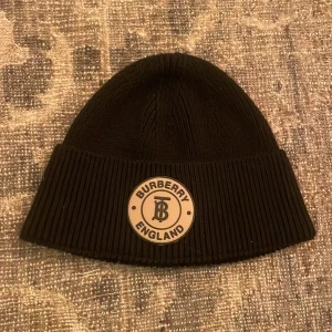 Svart ribbad mössa från Burberry - Säljer en snygg svart ribbad mössa från Burberry i 100% bomull. Den har en cool beige patch med Burberry-loggan framtill. Perfekt för att hålla sig varm och stilren under höst och vinter. Passar både till vardags och lite finare tillfällen.