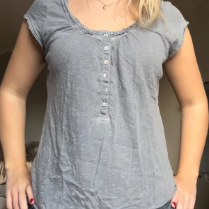 Supersöt grå topp med knappar - Säljer en grå topp i mjukt material med korta ärmar och knappar framtill. Den har en avslappnad passform och en fin detalj vid halsringningen. Perfekt för vardagsbruk!