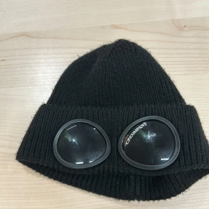 Svart mössa med glasögondetalj från C.P. Company - Säljer en cool svart mössa från C.P. Company med unika glasögondetaljer framtill. Den är ribbad och gjord i bomull, perfekt för att hålla värmen under höst och vinter. Mössan är i mycket bra skick och passar perfekt för en stilren look.
