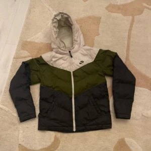 Flerfärgad Jacka från Nike - Säljer en snygg och varm polyester jacka från Nike i storlek XL KIDS, ungefär S i vanlig storlek. Jackan har en färgkombination av beige, grön och svart med en dragkedja framtill. Perfekt för kalla dagar med sin långa ärmar och huva.
