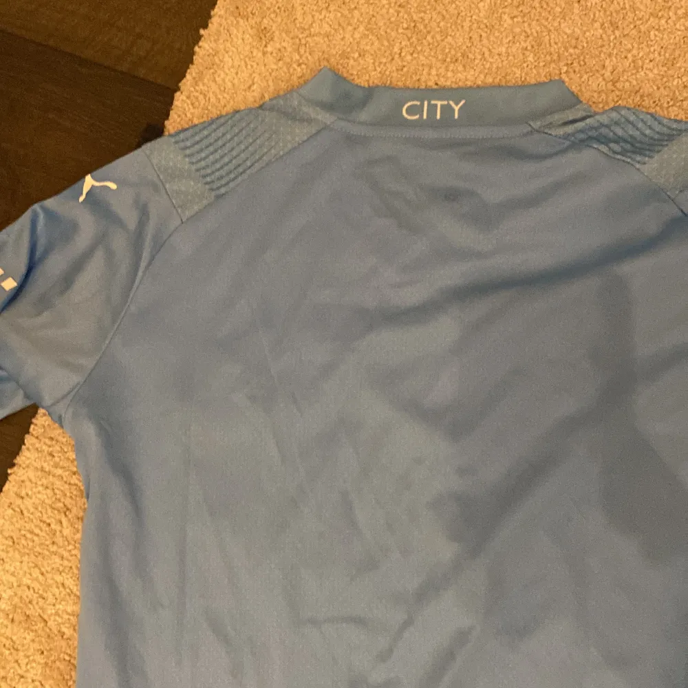 Säljer en ljusblå Manchester City fotbollströja från Puma. Den har korta ärmar och klubbens emblem på bröstet. Tröjan är i mycket bra skick och har ett snyggt mönster med ränder. Perfekt för alla City-fans! 💙. T-shirts.