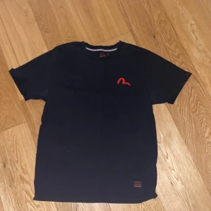 Svart t-shirt från Evisu - Säljer en svart t-shirt från Evisu i storlek L. Den har en cool röd logga på bröstet och ett stort, färgglatt mönster på ryggen med Evisu-tryck. Perfekt för en avslappnad stil eller streetwear-look. T-shirten är i bra skick och gjord av bomull.
