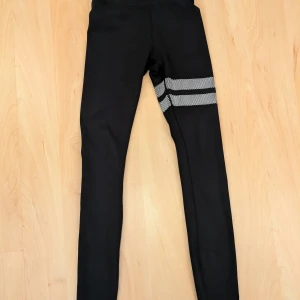 Svarta leggings från Stronger - Snygga svarta leggings från Stronger med vita randiga detaljer runt låret. Perfekta för träning eller en avslappnad dag. De är stretchiga och bekväma med hög midja för extra stöd. Kan bli lite tajt runt midjan