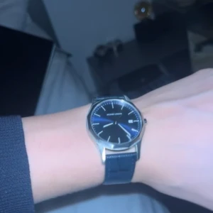 Elegant herrklocka med blå läderarmband - Snygg och stilren herrklocka med en silverfärgad boett och blå urtavla. Klockan har ett klassiskt utseende med ett svart läderarmband som ger en tidlös känsla. Perfekt för både vardag och festliga tillfällen.